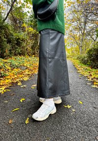 Black leather maxi skirt with a smooth texture, paired with white trainers. A green knitted sweater and a small black shoulder bag are visible.
