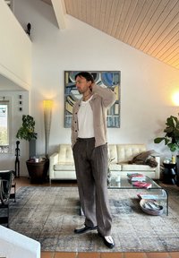 Beige cardigan over a hvit t-skjorte, grå rutete bukser, svarte sko med hvite detaljer. Moderne stue med et glass kaffebord og kunst.