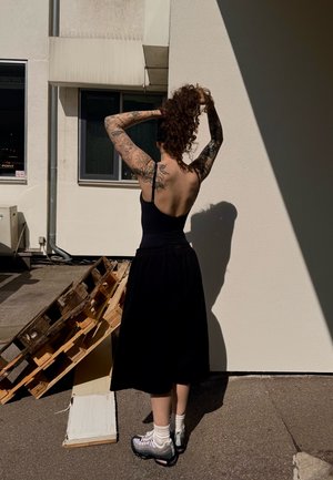 Top negro sin mangas y falda larga negra, adornada con tatuajes. Lleva calcetas blancas y zapatillas gris-negras. El fondo presenta un palé de madera.