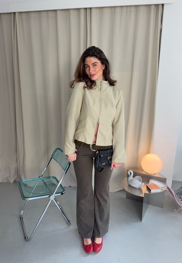 Une femme pose à côté d’une chaise pliante transparente. Elle porte une veste couleur crème, un pantalon foncé, un sac noir en bandoulière et des chaussures plates rouges. Une sphère lumineuse et des décorations sont posées sur une table à proximité.