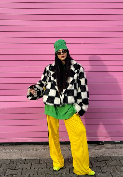 Mujer con chaqueta de piel a cuadros en blanco y negro, gorro verde, sudadera, pantalones amarillos, zapatos neón y gafas de sol, de pie frente a una pared rosa.