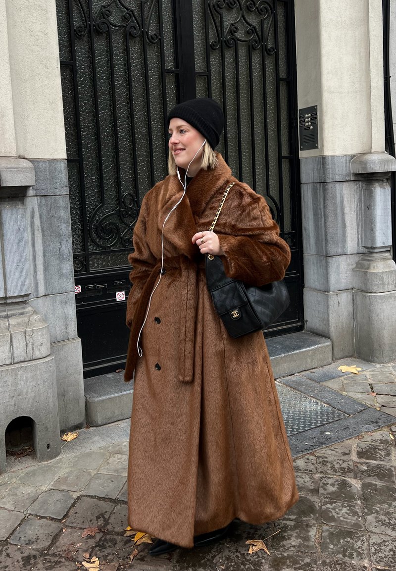 Femme portant un long manteau en fourrure marron et un bonnet noir, tenant un sac matelassé noir avec une bandoulière en chaîne, debout sur une rue pavée près d'une grille en fer noir.