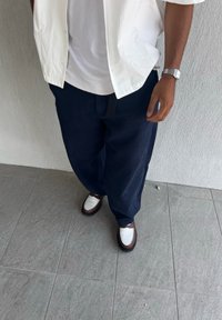 Pantalons larges bleu marine, T-shirt blanc et chemise blanche à boutons ; des chaussures brunes et blanches avec un design texturé complètent la tenue.