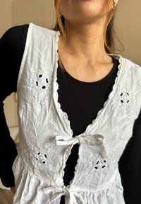 Fehér hímzett mellény virágmintákkal, hullámos szélekkel és megkötős záródással. Fekete hosszúujjú felsőre van rétegezve.