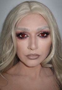 Nahaufnahme eines Gesichts mit langen, welligen platinblonden Haaren. Betont rote Augen, definierte Augenbrauen und neutrale Lippenfarbe. Dramatisches Augen-Make-up.