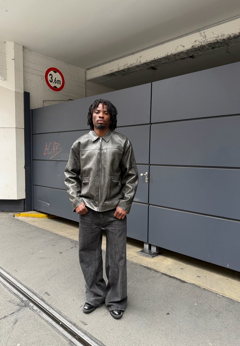 Homme aux dreadlocks portant une veste en cuir grise et un pantalon ample foncé, debout dans une rue près d'une porte de garage grise et d'un panneau indiquant une hauteur de 3,6 m.