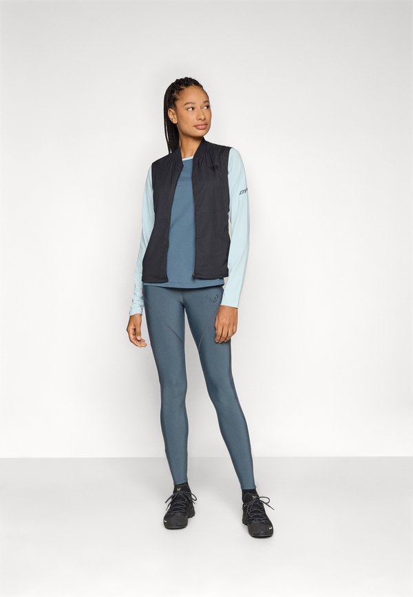 Čierny bezrukávový sveter cez svetlomodrý top s dlhými rukávmi, doplnený morskými legínami. Má hladký materiál a športový dizajn. Čierne tenisky.