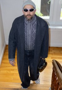 Manteau en laine gris, pull en tricot à motifs, jean foncé et baskets blanches. Accessoire : petit sac à main noir. Lunettes de soleil oversize et bonnet.