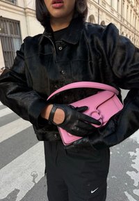 Schwarze Kunstpelzjacke, schwarze Lederhandschuhe, die eine kleine rosafarbene Lederhandtasche mit einem runden Griff und kreisförmigen Beschlägen halten.