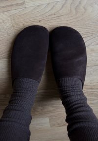 Πόδια που φορούν σκούρα καφέ σουέντ παντόφλες και παχύ ριγέ γκρι καλτσάκια πάνω σε ανοιχτό ξύλινο πάτωμα.