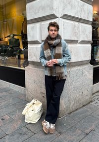 Veste en denim, écharpe à carreaux marron et gris, pantalon noir, baskets marron et un sac crème. Un homme se tient près d'un mur en pierre près d'une vitrine.