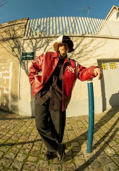 Chaqueta de cuero roja con detalles blancos y logo "NY", camiseta negra, pantalones holgados negros y zapatos negros. Gorro de lana con rayas blancas y negras.