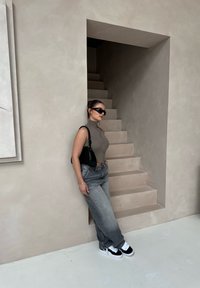 Grå ribbet ærmeløs top med høj hals, parret med løse grå jeans og sorte sneakers. Minimalistisk trappebaggrund i neutrale nuancer.