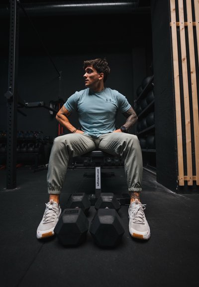 Camiseta deportiva azul claro de manga corta, pantalones joggers verde oliva y zapatillas grises. Sentado en un banco con pesas negras en el suelo del gimnasio.