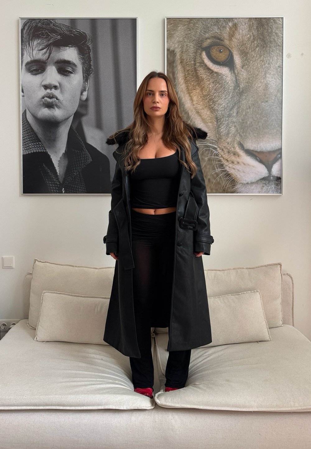 Manteau noir en cuir à double boutonnage avec col en fourrure, porté sur un haut noir et des leggings ; personne debout sur un canapé de couleur claire, chaussures rouges visibles.