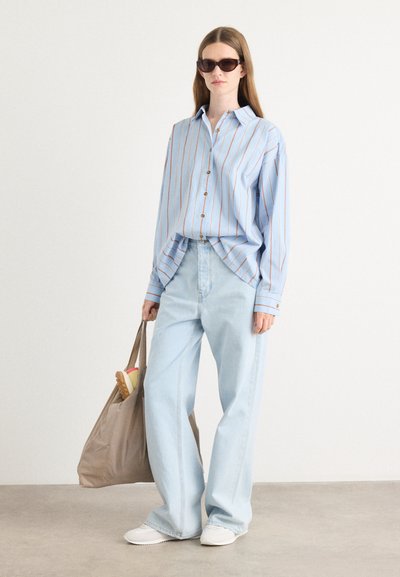 Camisa de rayas azul claro con líneas marrones, con botones en la parte delantera, combinada con jeans azul claro de corte relajado y zapatillas blancas. Sostiene un bolso tote marrón.