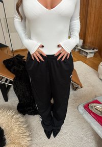 Body blanc à manches longues et joggeurs noirs avec des bandes blanches sur les côtés, assortis à des talons noirs à bout pointu. Tapis en peluche texturé en dessous.