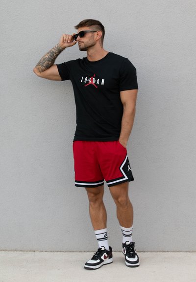 Camiseta negra con gráfico blanco y rojo, pantalones cortos rojos con acentos negros, zapatillas negras y calcetines a rayas; modelo usando gafas de sol.