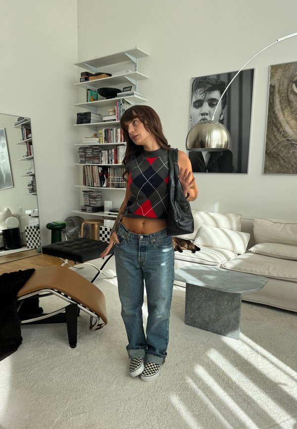 Joven mujer con top sin mangas de rombos, vaqueros y zapatos a cuadros está de pie en una sala de estar moderna con un sofá blanco, estanterías y una gran obra de arte en la pared.