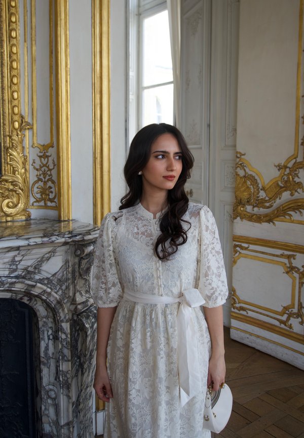 Woman in white lace dress with puff sleeves and bow belt stands near ornate marble fireplace and gold-trimmed walls, holding a white clutch.