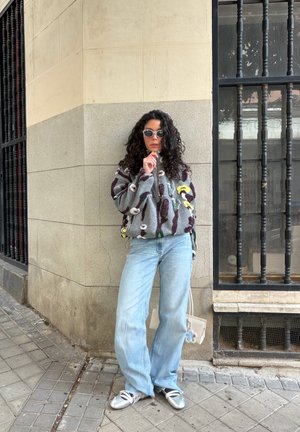 Mujer con cabello rizado oscuro, usando gafas de sol, chaqueta estampada, jeans azul claro, zapatos plateados y sosteniendo un bolso blanco mientras está de pie en una acera.