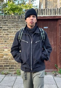 Jacket preta com fecho éclair e gola alta, combinada com calças cargo verde-azeitona e um gorro preto. O sujeito tem uma mochila com alças verdes.