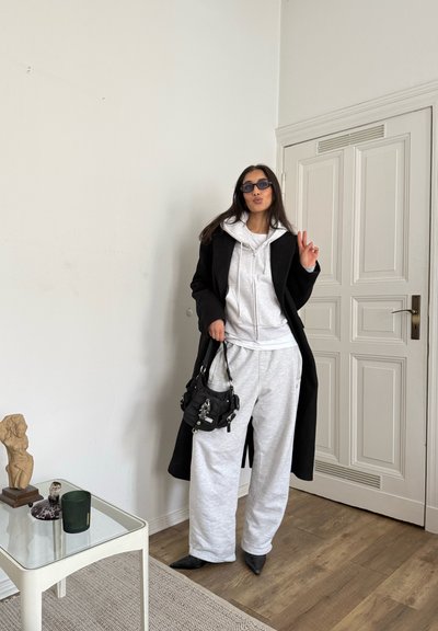 Abrigo negro sobre una sudadera con capucha blanca y pantalones de chándal grises, combinado con zapatos negros puntiagudos y un pequeño bolso negro. El fondo presenta una pared blanca y suelo de madera.