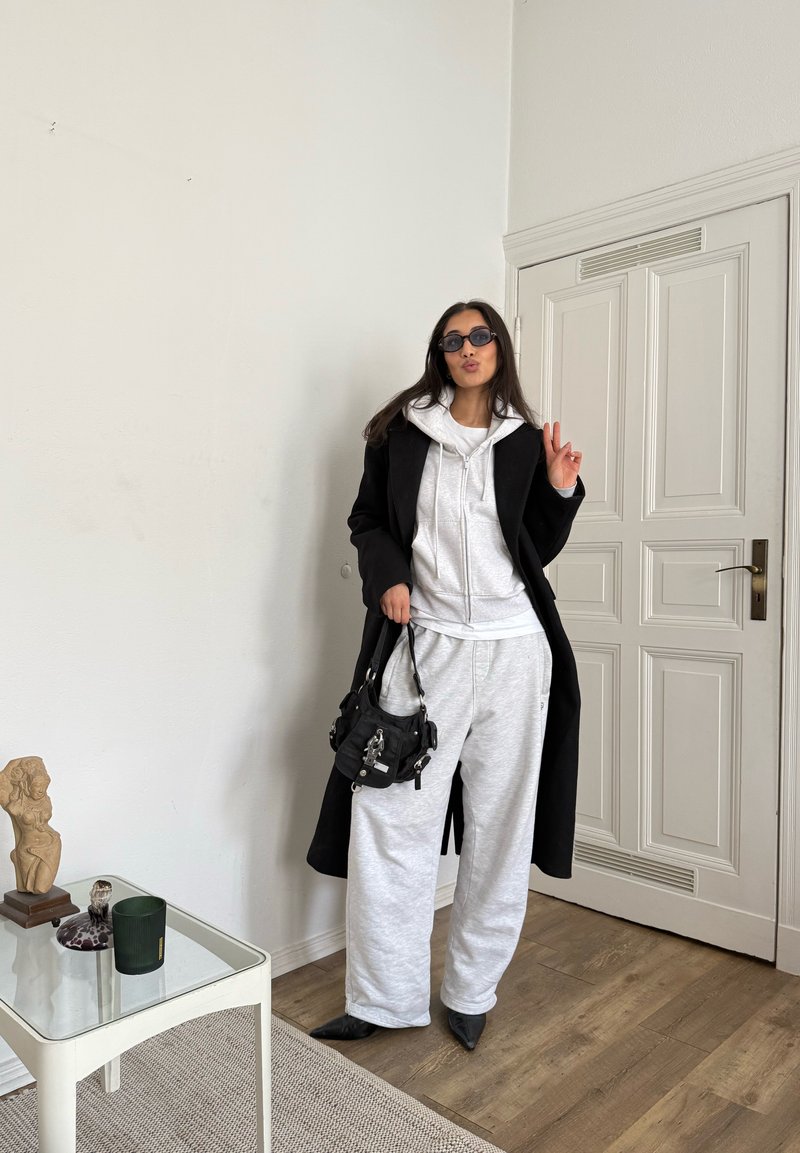 Black coat worn over a white hoodie and grey sweatpants, paired with pointed black shoes and a small black handbag. The background features a white wall and wooden flooring.