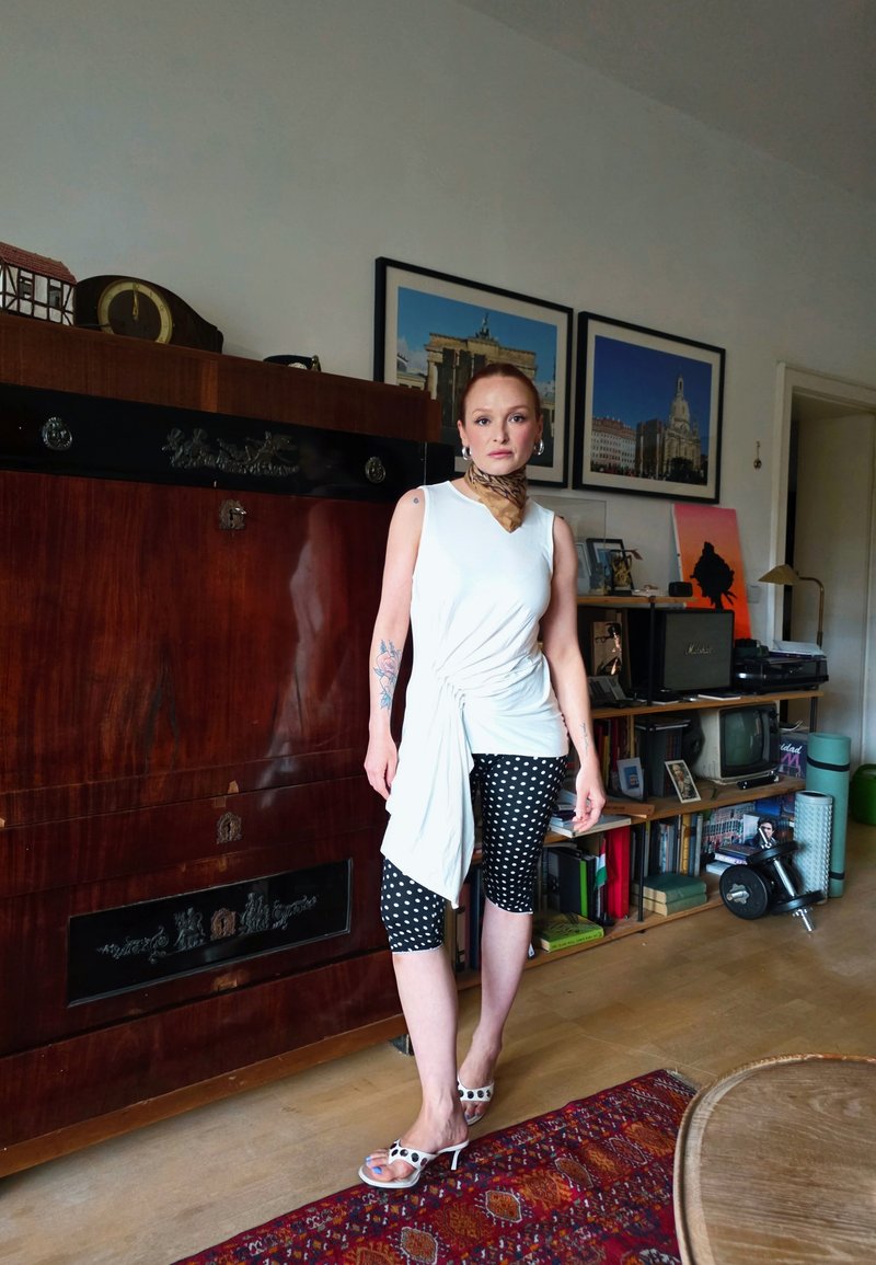Femme avec des tatouages portant un haut blanc sans manches, des leggings à pois et des talons, debout dans le salon avec une armoire vintage et des photos encadrées.