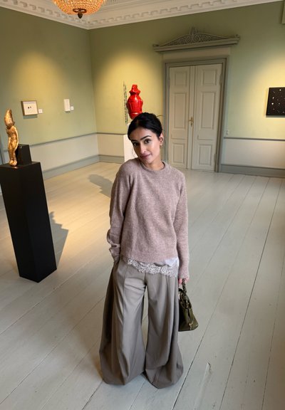 Joven mujer con suéter beige y pantalones anchos sosteniendo un bolso verde está en una galería de arte con esculturas y pinturas.
