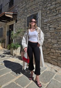 Manteau beige par-dessus un débardeur blanc, pantalon noir court et tongs noires ; tenant un sac à main rouge ; mur de pierre en arrière-plan.