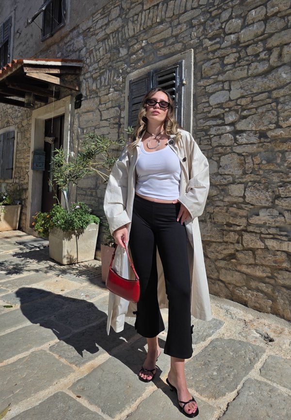 Beige trench coat over a white tank top, black cropped pants, and black flip-flops; holding a red handbag; stone wall background.
