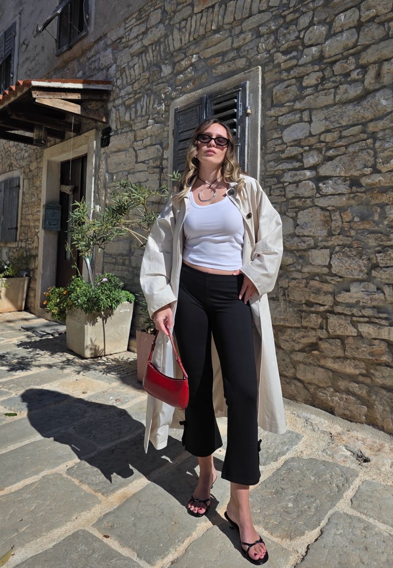 Manteau beige par-dessus un débardeur blanc, pantalon noir court et tongs noires ; tenant un sac à main rouge ; mur en pierre en arrière-plan.