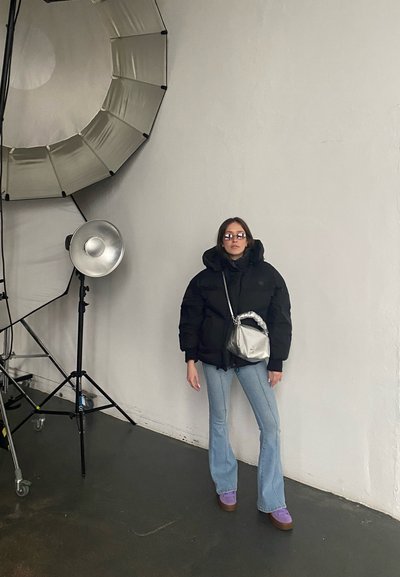 Chaqueta de plumas negra, jeans acampanados azul claro, bolso metálico plateado, zapatos morados, de pie contra una pared lisa con iluminación de estudio.