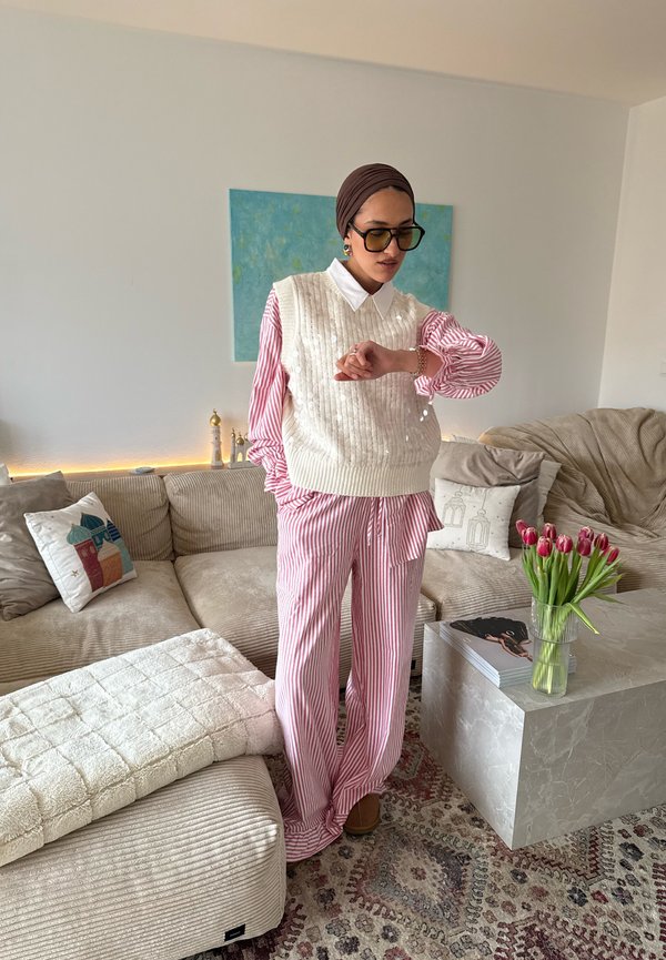 Femme vêtue d'une tenue rayée rose et blanche avec un gilet en tricot blanc et un foulard marron, regardant sa montre, debout dans un salon chaleureux avec un canapé beige et des tulipes.