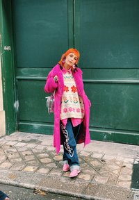 Abrigo de peluche rosa, suéter de punto colorido con patrón floral, superpuesto sobre una camisa rosa. Jeans azul oscuro con texto blanco, zapatos rosas.