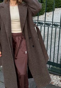 Brun ullkåpe med store slag og to knapper, lagd over en beige topp og mørkebrune satengbukser med hvitt logo og striper.