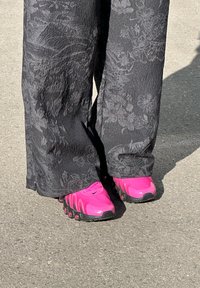 Calçados atleticos rosa com um upper de malha texturizada, sola preta e pontas arredondadas, combinados com calças largas pretas com padrão floral.