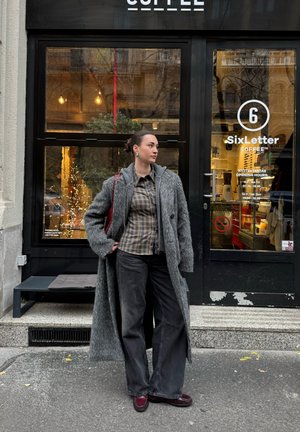 Abrigo gris de gran tamaño con acabado texturizado, camisa estampada, pantalones negros de pierna ancha y zapatos burdeos; ambientado fuera de una cafetería.
