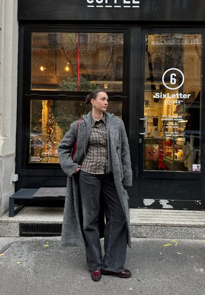 Abrigo gris de gran tamaño con acabado texturizado, camisa estampada, pantalones negros de pierna ancha y zapatos burdeos; ambientado fuera de una cafetería.