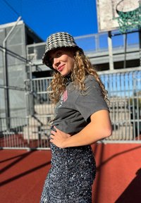 Fekete-fehér houndstooth mintás vödörsapka, szürke grafikus póló, fényes ezüst flitteres szoknya, piros kosárlabdapályán állva, látható kosárlabda gyűrűvel.