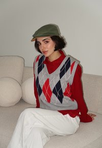 Γκρι αργυλωτό γιλέκο πάνω από κόκκινο πουκάμισο με γιακά, συνδυασμένο με λευκά παντελόνια. Ένα πράσινο καπέλο με καφέ γείσο ολοκληρώνει την εμφάνιση. Στον φόντο, καναπές με πιο απαλή υφή.