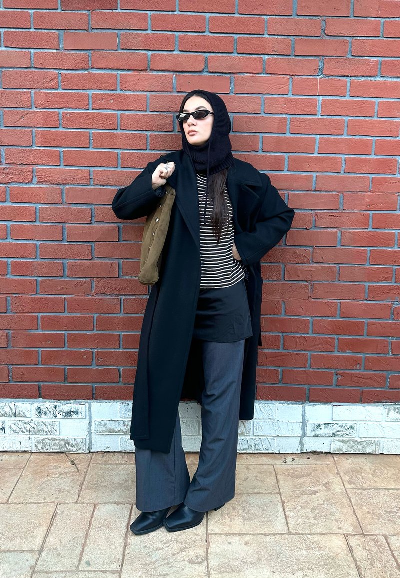 Manteau noir long par-dessus un haut rayé, pantalon gris évasé, chaussures noires et sac vert ; debout contre un mur en briques.