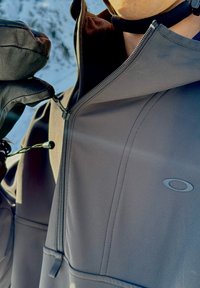 Veste noire à texture lisse et mate, dotée d'une fermeture éclair à l'avant et d'un logo. Une main gantée ajuste la fermeture éclair.