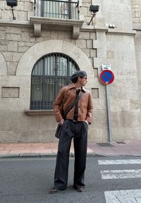 Casaco curto em pele castanha, camisola preta, calças largas escuras e boné cinzento. Visual casual combinado com uma mala de ombro. Fundo com edifício de pedra.