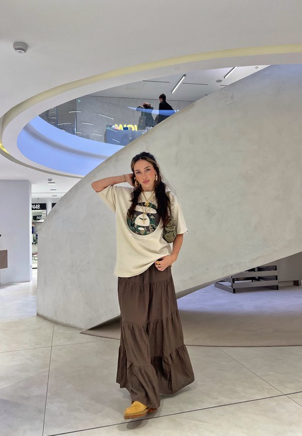 Beige t-paita pyöreällä graafisella printillä, kerroksellinen ruskea maksihame ja keltaiset liukupohjaiset kengät. Sisätila, jossa on kaareva valkoinen seinä ja moderni design.