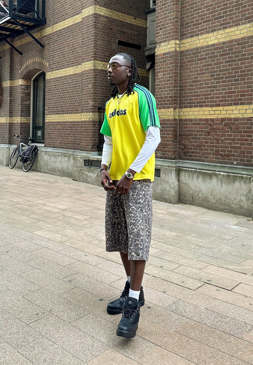 Sportovní oblečení obsahující žlutozelený pruhovaný dres s logem "adidas", vzorované šortky a černé boty. Městské prostředí s cihlovou zdí.