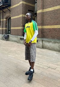 Sportovní oblečení obsahující žlutozelený pruhovaný dres s logem "adidas", vzorované šortky a černé boty. Městské prostředí s cihlovou zdí.