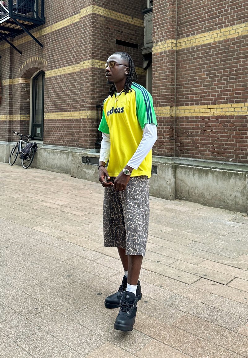 Idrottskläder som visar en gul och grön randig tröja med "adidas"-logotyp, mönstrade shorts och svarta stövlar. Gatumiljö med tegelvägg.