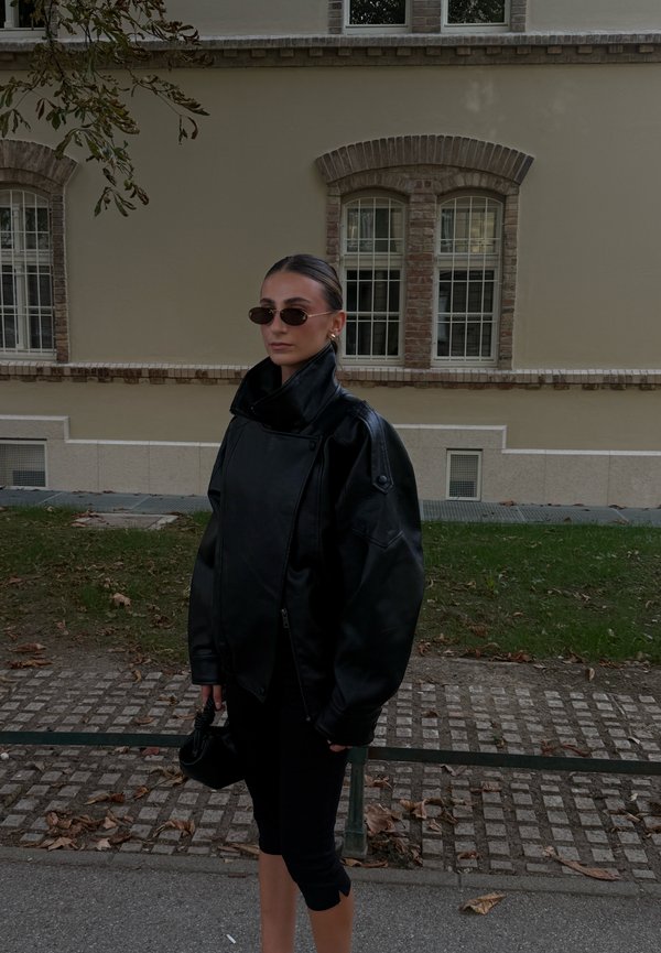 Femme portant une veste en cuir noire oversize, une jupe noire, de petites lunettes de soleil noires et tenant un petit sac à main noir, debout à l'extérieur.